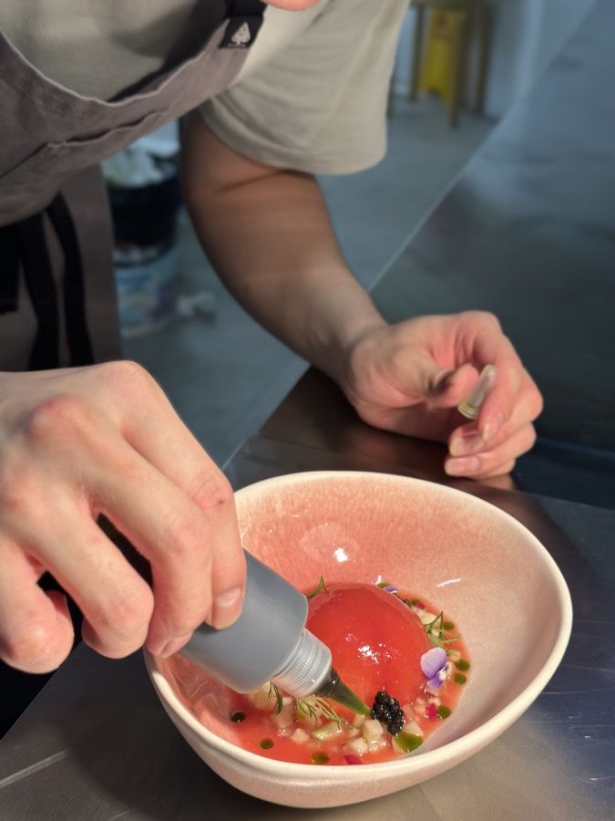 A chef finishing a pink bowl of tomato, caviar and herbs with a squeeze bottle