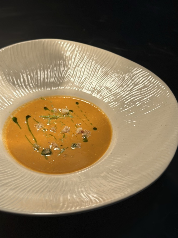 An orange bisque with herb oil in a wide, textured white bowl